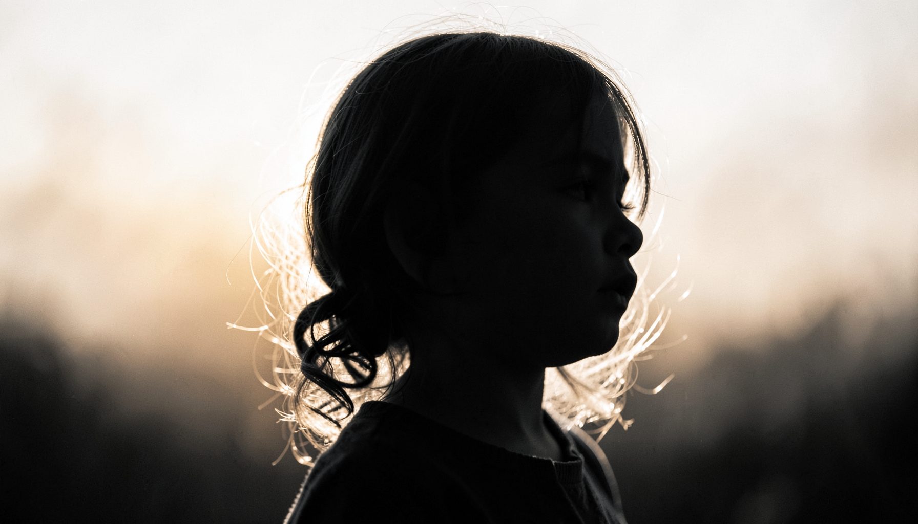 A little girl's silhouette against soft golden light, delicate curls catching the glow, tender black-and-white portrait mood