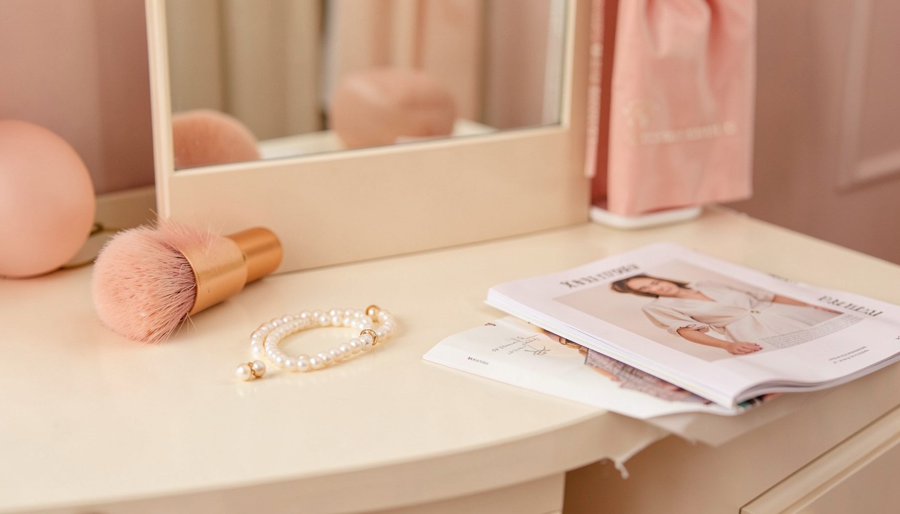A softly lit vanity table with a pink blush brush, a pearl bracelet, and a folded fashion magazine, warm rose and cream tones