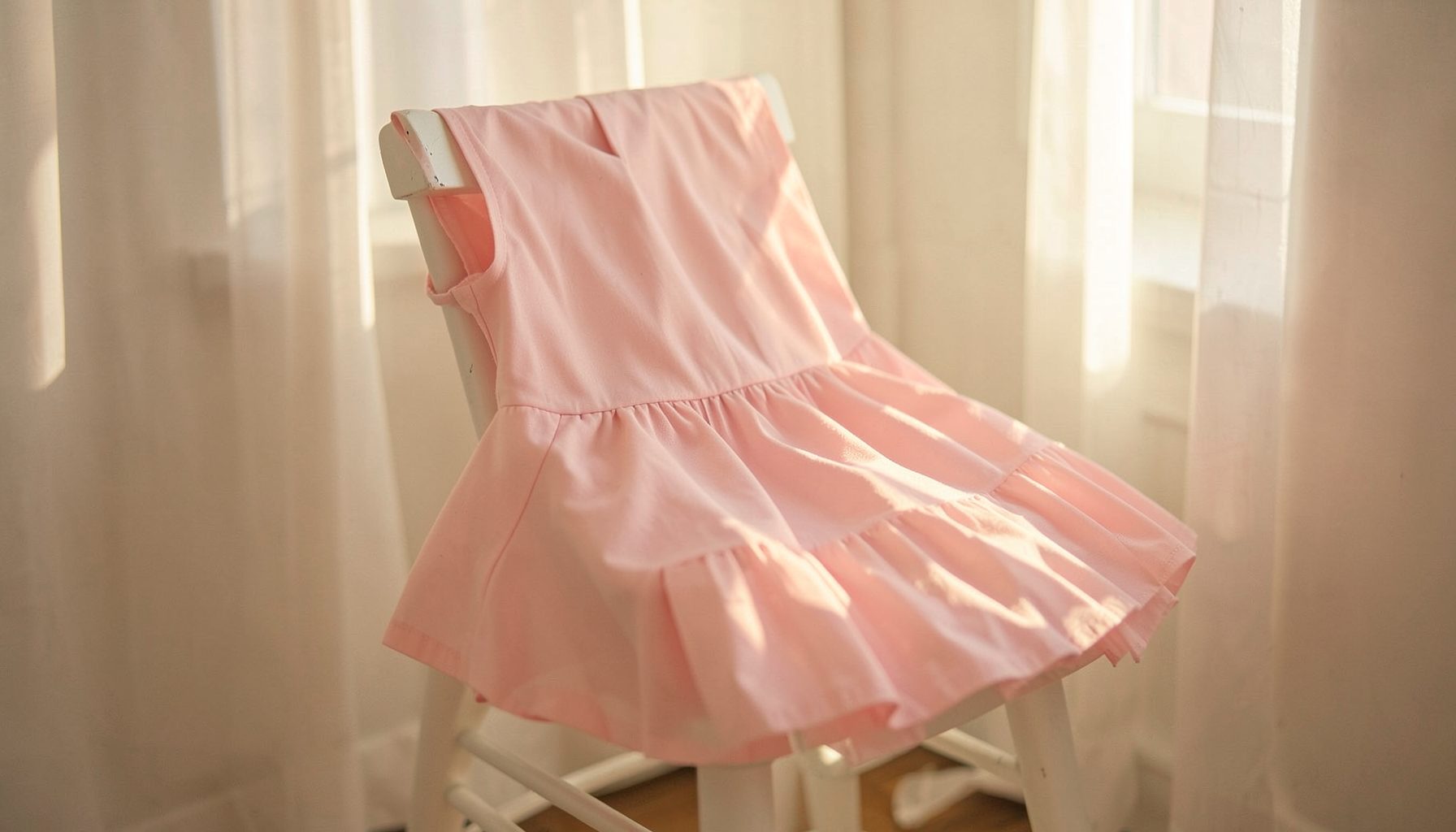 Soft pink tutu skirt draped over a white wooden chair, warm sunlight streaming through sheer curtains, gentle and dreamy atmosphere